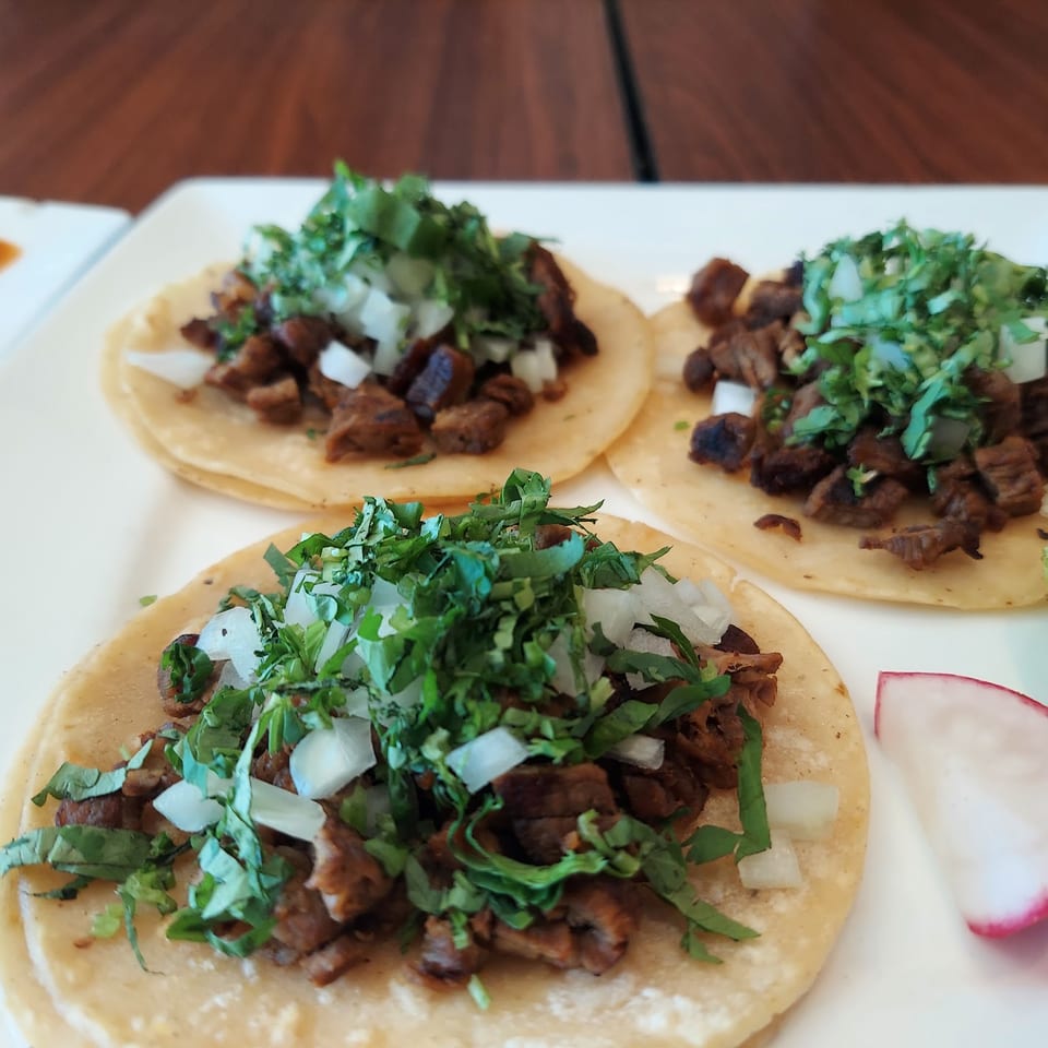 Three street tacos with meat, cilantro, and onions, served with lime and radish slices.
