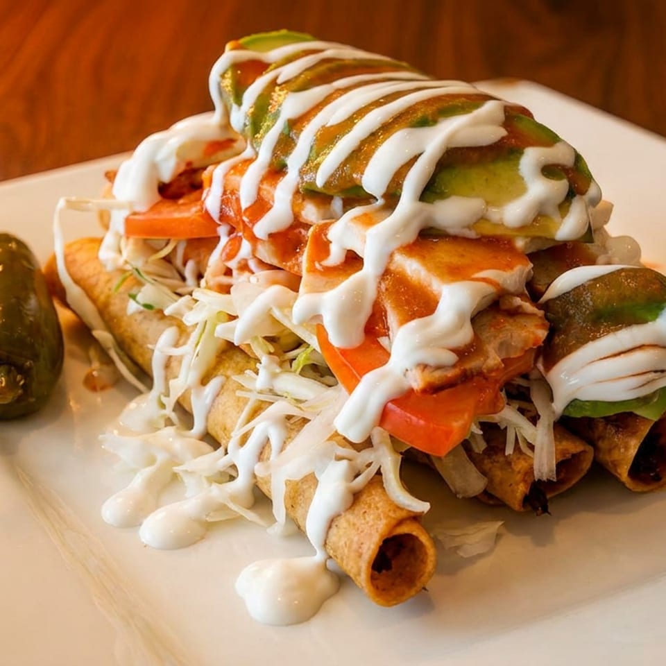 Crispy taquitos topped with shredded cabbage, diced tomatoes, avocado, and drizzled with crema.