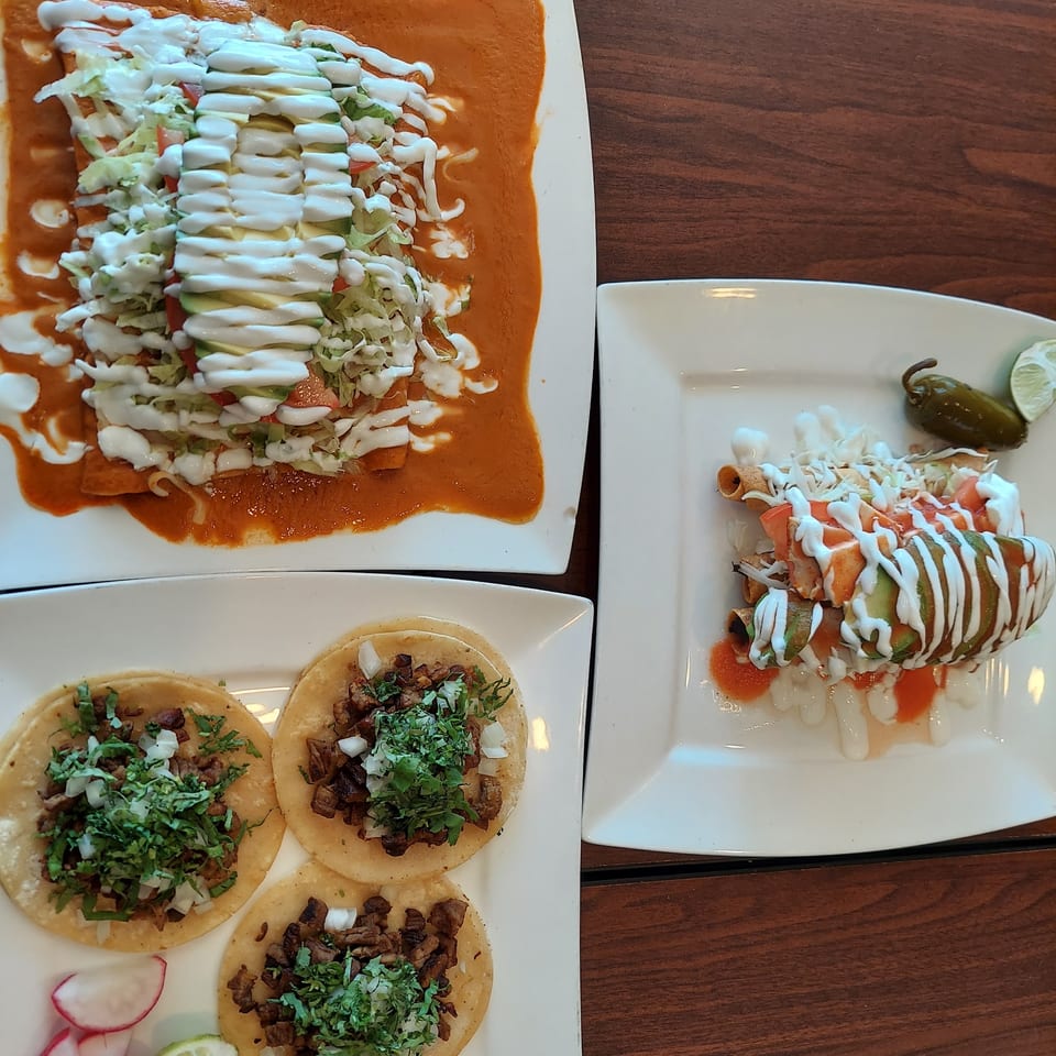 Three plates of Mexican food, including enchiladas, flautas, and street tacos, on a wooden table.